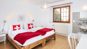 A bedroom with a wooden bed, white bedding, a red blanket, a window with a view, a white radiator, and a white sink in the corner.