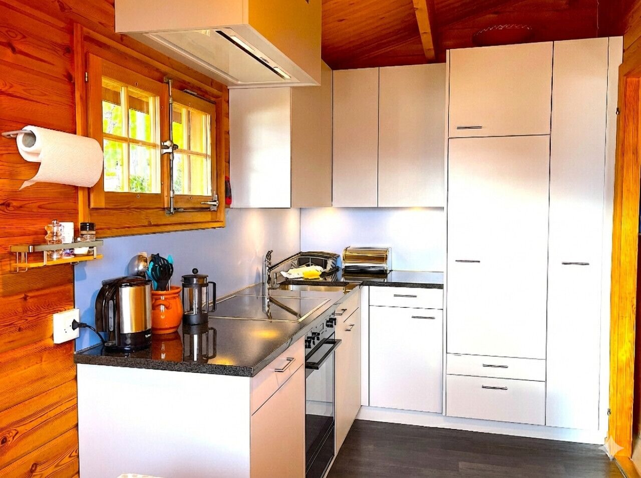 A compact kitchen with wooden walls, white cabinets, a countertop with a kettle, toaster, and coffee pot.
