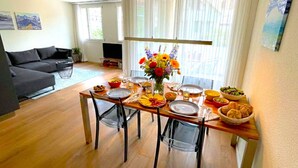 A dining area with a wooden table set for four, surrounded by chairs. A vase with flowers is placed in the center.