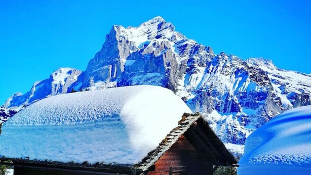 A wooden cabin with a snow-covered roof, surrounded by snow and mountains in the background.