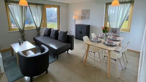 A modern living room with a sofa, coffee table, and dining area with chairs and a table set for two. - Concierge, Lake Thun/Alps Scenic Views by SwissHut (Sigriswil)