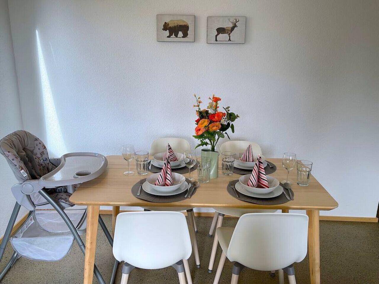 A dining table set for four with white chairs, a baby high chair, and a vase of flowers.