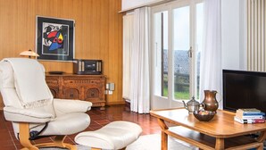 A living room with a white armchair, a wooden coffee table, a television, and a view of the outdoors through large windows.