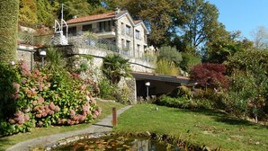 A stone house with a balcony, surrounded by lush greenery and a small pond.