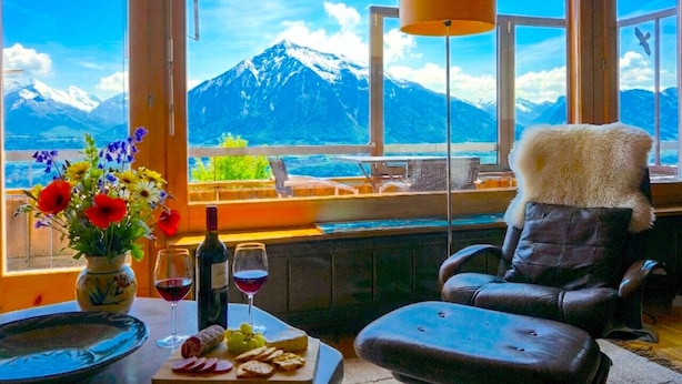 A room with a view of a mountain, a table with food and drinks, a chair, and a vase with flowers.