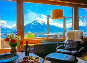 Una habitación con vista a la montaña, una mesa con comida y bebidas, una silla y un florero con flores.
