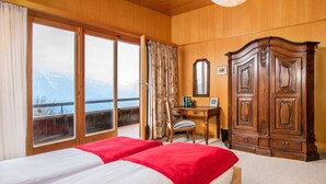 A hotel room with two beds, a wooden wardrobe, a desk with a chair, and a large window offering a mountain view.