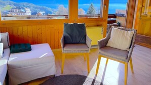 A cozy room with a bed, two wicker chairs, and a view of mountains through large windows.