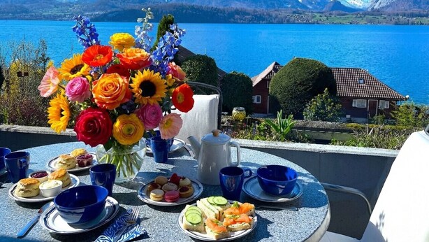 A table set for breakfast with a mountain view, a bouquet of flowers, and a scenic lake.