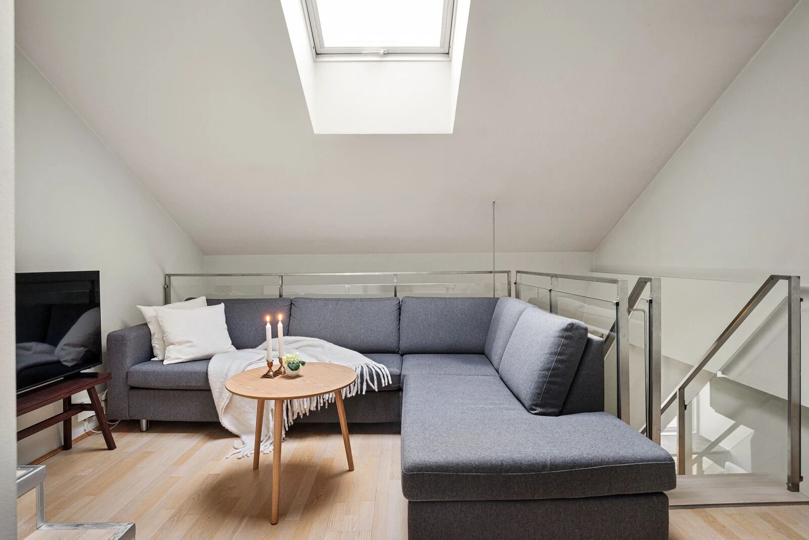 A modern loft apartment with a grey sectional sofa, a small round wooden table with candles, and a TV on a wooden stand.