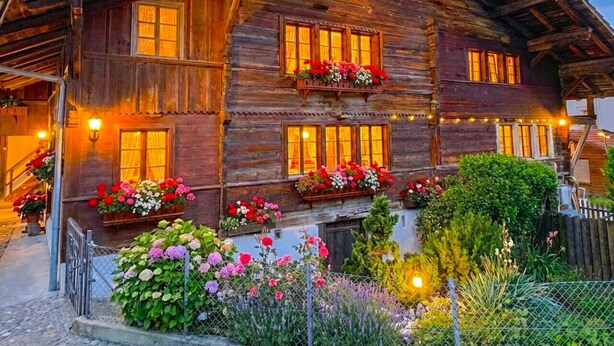 A rustic wooden chalet with multiple windows, a balcony with flower boxes, and a well-lit entrance.