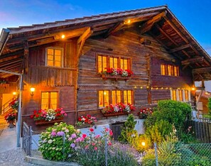 Un chalet rústico de madera con múltiples ventanas, un balcón con macetas de flores y una entrada bien iluminada.