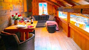 A cozy attic room with a wooden interior, a dining table set with wine glasses, a sofa, and a view of a stone-lined water feature through a window.