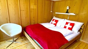 Una habitación con paneles de madera, una cama, una silla blanca y una mesita de noche con una lámpara.