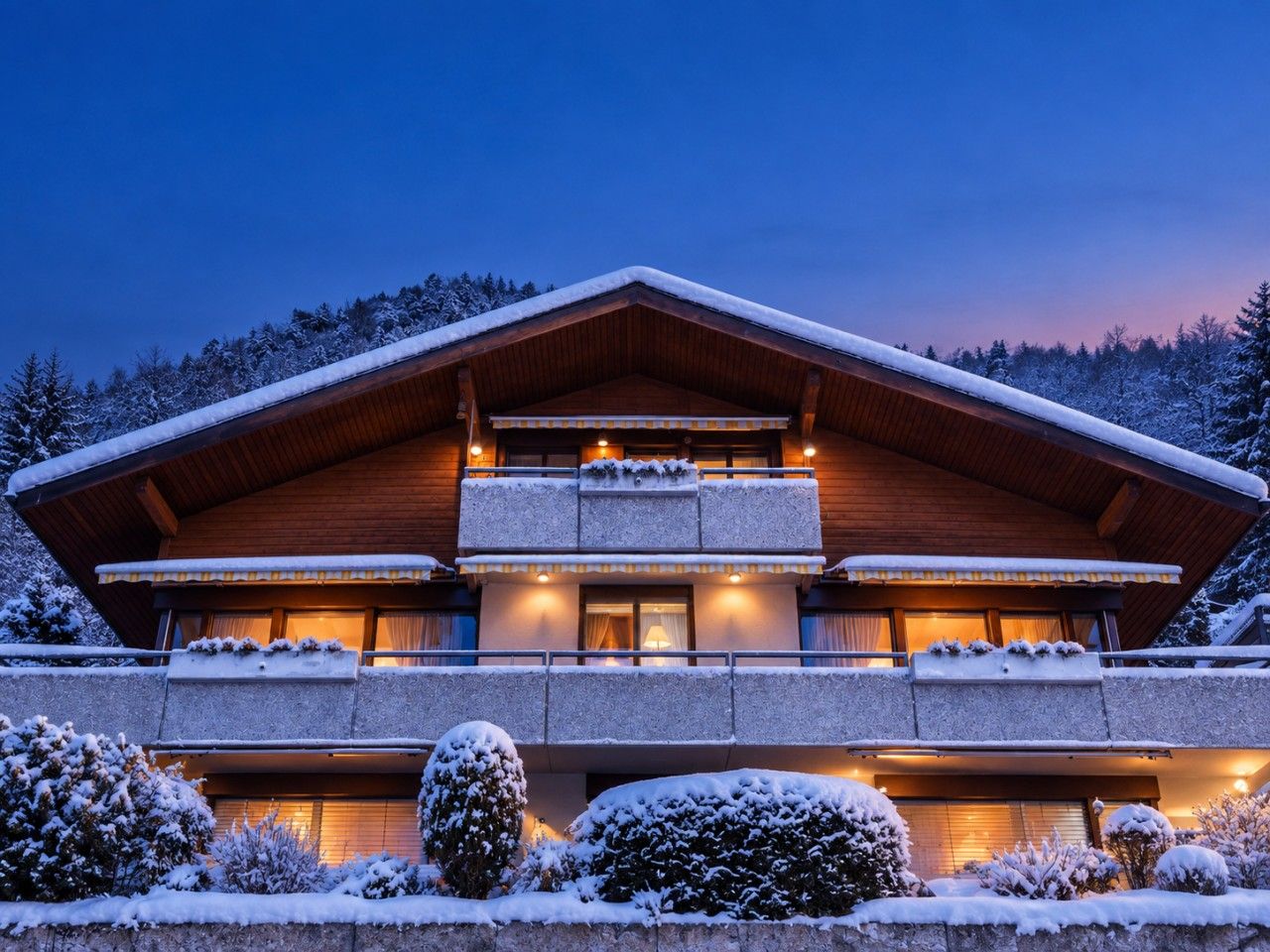 Un chalet en bois à deux étages, doté d’un balcon et entouré d’arbres couverts de neige.