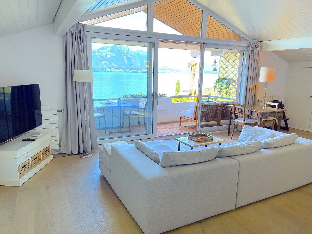 Ein modernes Wohnzimmer mit einer großen Couch, einem Flachbildfernseher und einer Schiebetür zu einem Essbereich im Freien mit Blick auf einen See und Berge.
