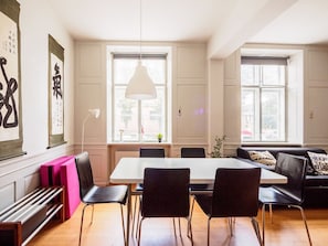 A spacious living area with a dining table, chairs, and a sofa. There are large windows providing natural light and a view outside.