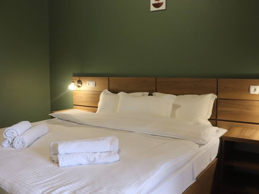 Cama bien hecha con sábanas blancas y toallas enrolladas, cabecera de madera y luces fijadas a la pared.