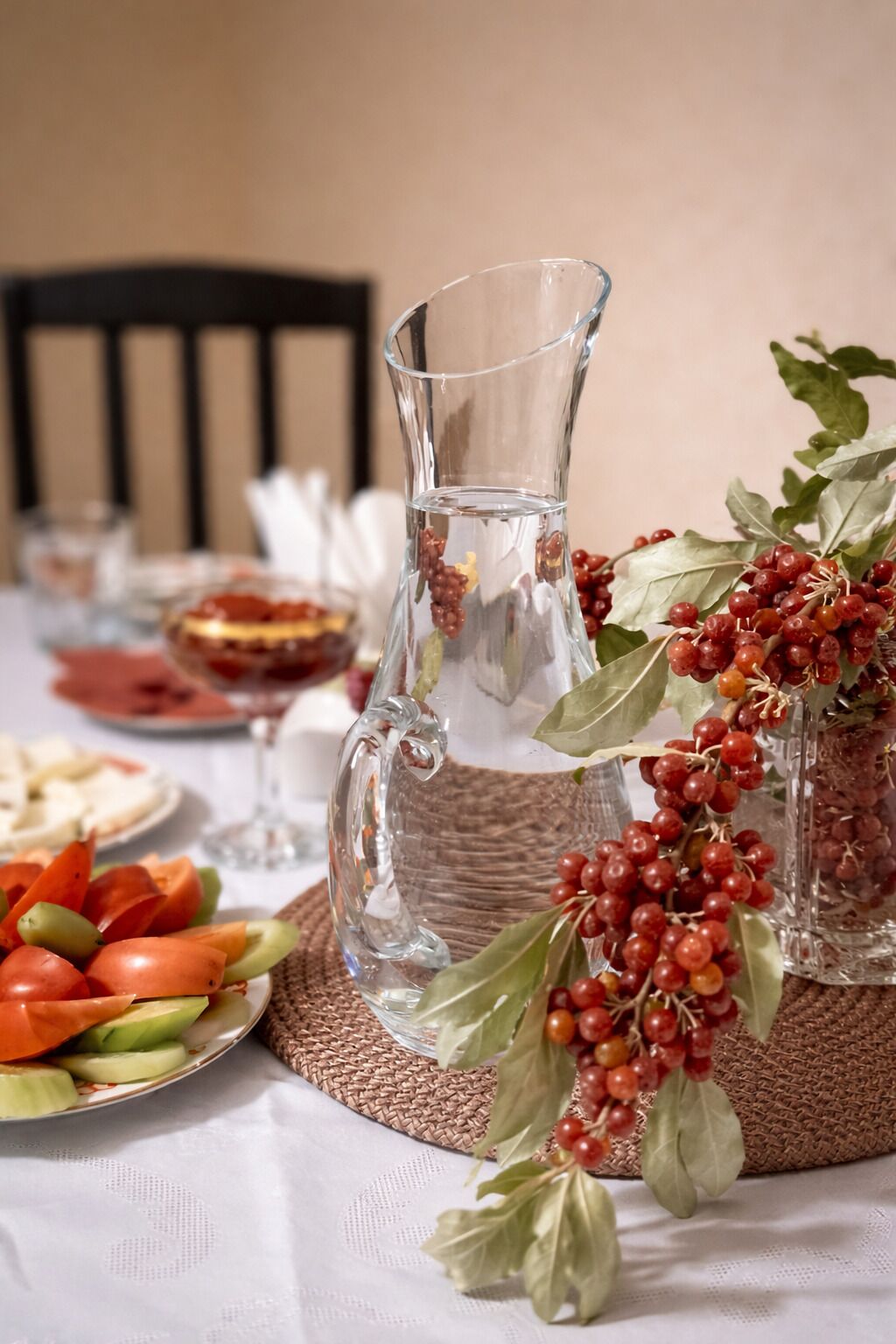 Una mesa dispuesta con una jarra de vidrio transparente, un plato de vegetales cortados y una decoración de bayas rojas y hojas verdes.