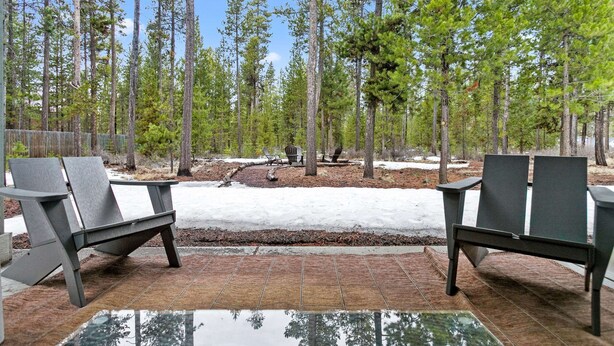 Un área de estar al aire libre con dos sillas Adirondack y una mesa de vidrio, rodeada por un bosque de altos pinos y suelo cubierto de nieve.