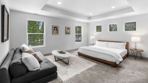 Un dormitorio moderno con una cama grande, un sofá, una mesita de centro redonda y varias ventanas que dejan entrar luz natural.