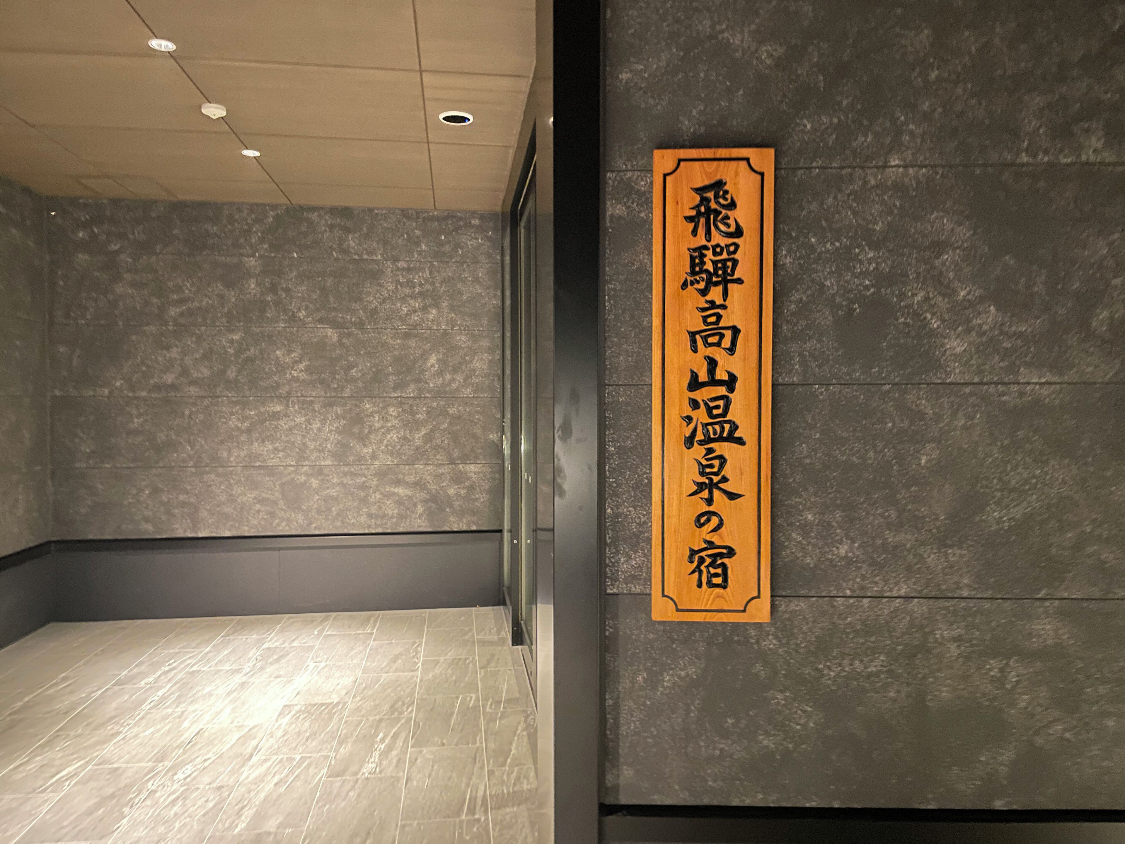 A hotel interior with a sign in Japanese on the wall, a tiled floor, and a ceiling with recessed lighting.