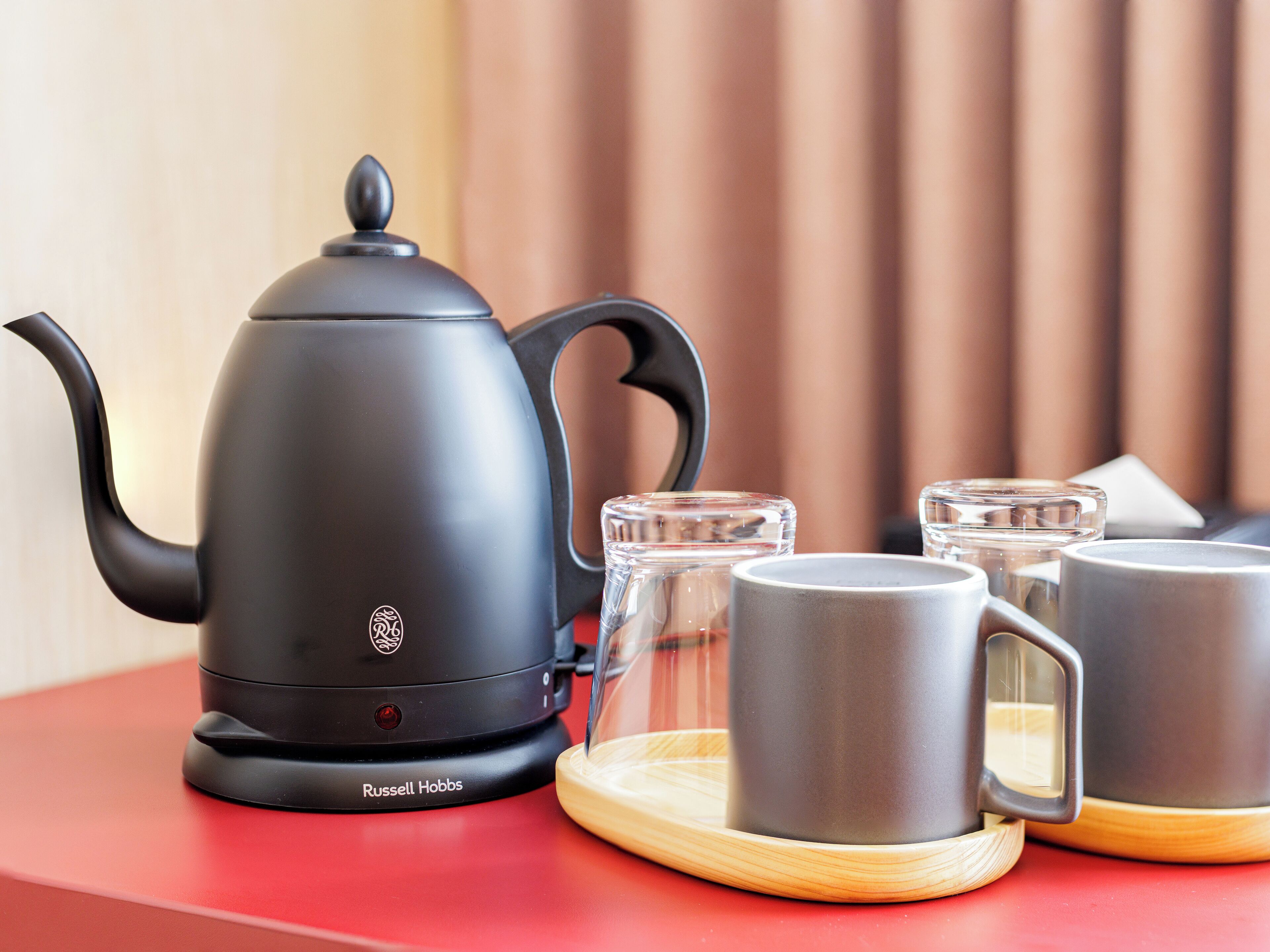 A black kettle, two clear glasses, and two grey mugs on a wooden coaster.
