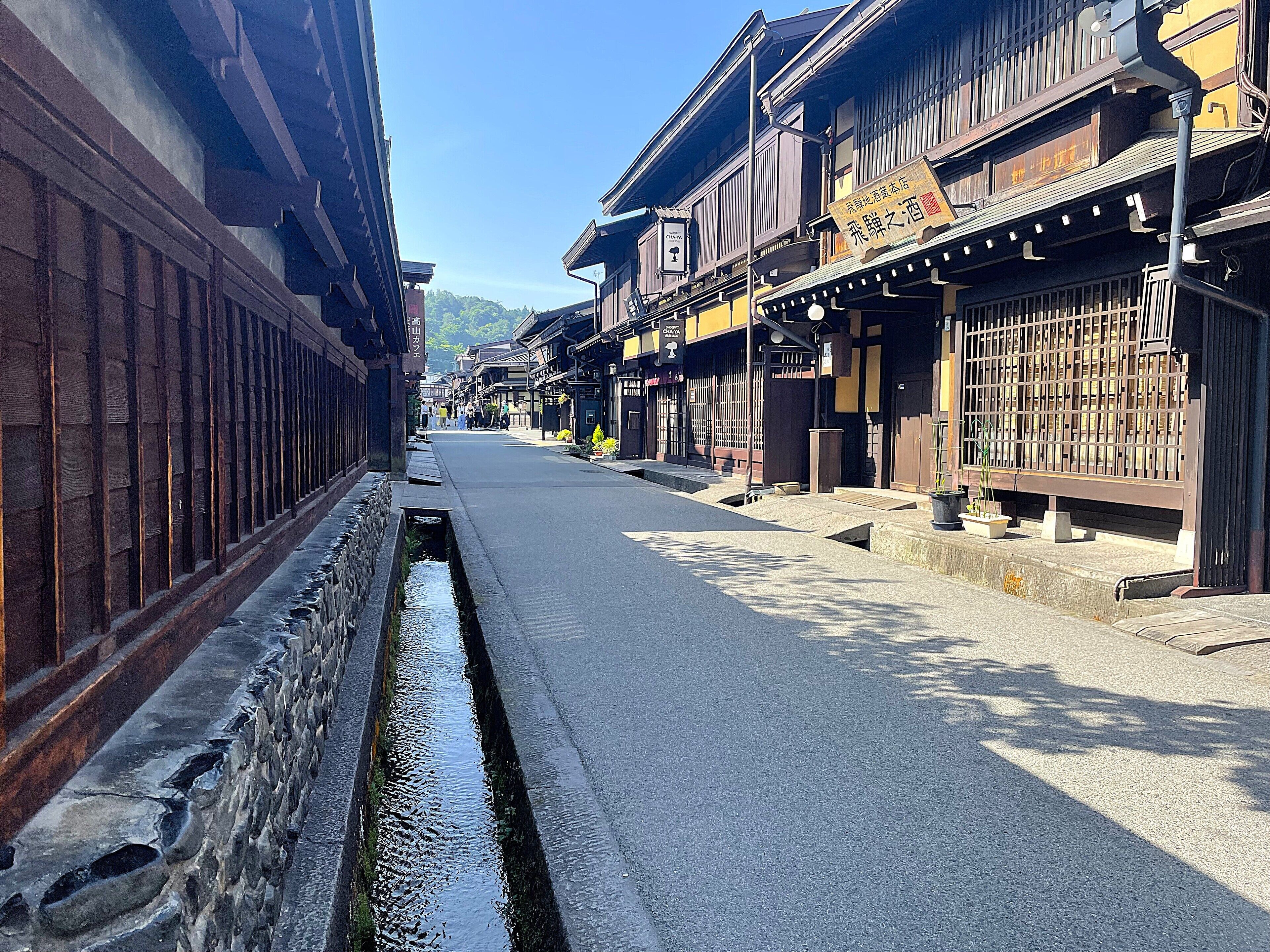 A narrow street with traditional wooden buildings, a stone water channel, and a clear sky.