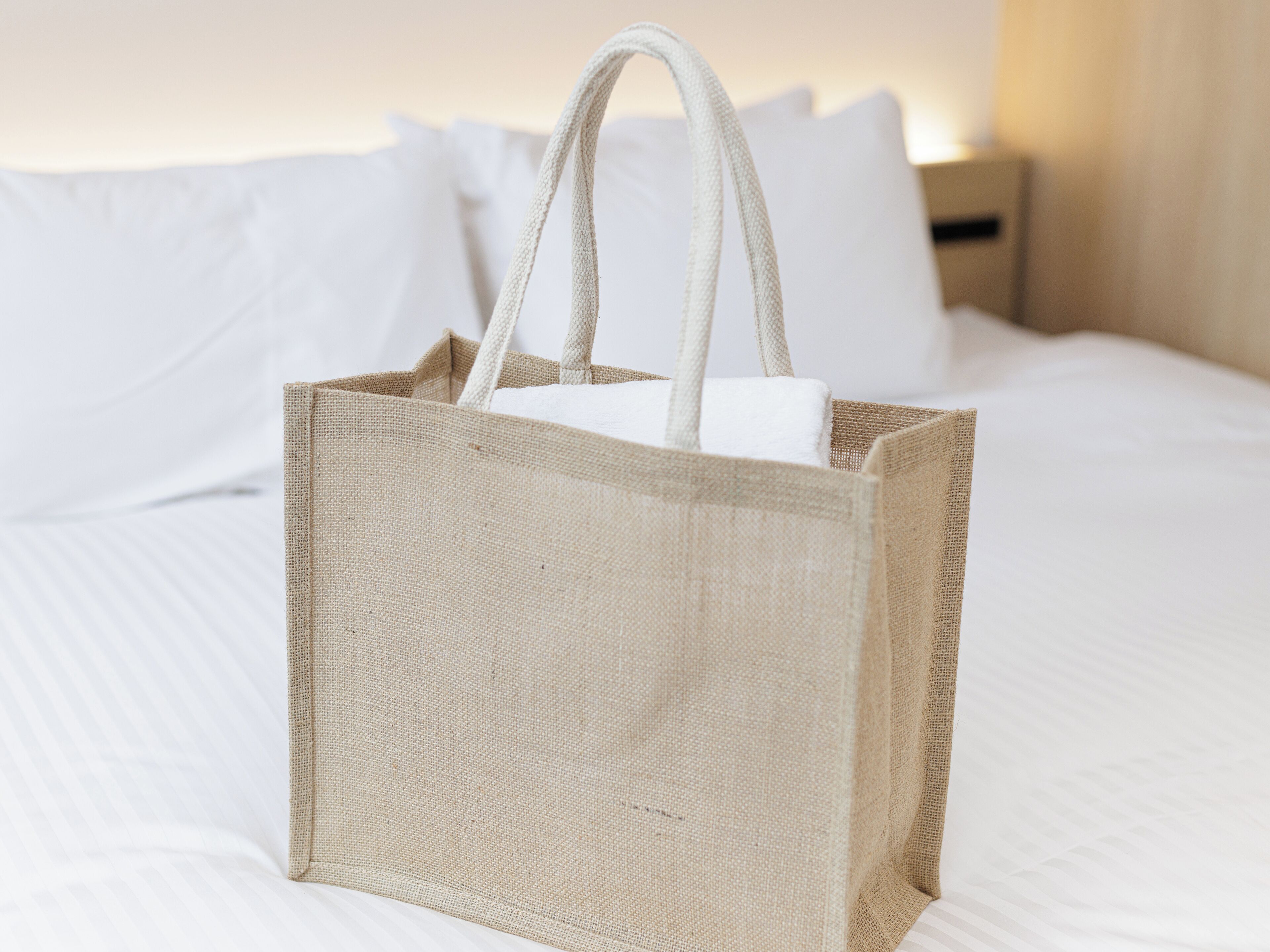 Eine große, beige gewebte Tragetasche mit Henkeln, platziert auf einem Bett mit weißer Bettwäsche und Kissen.