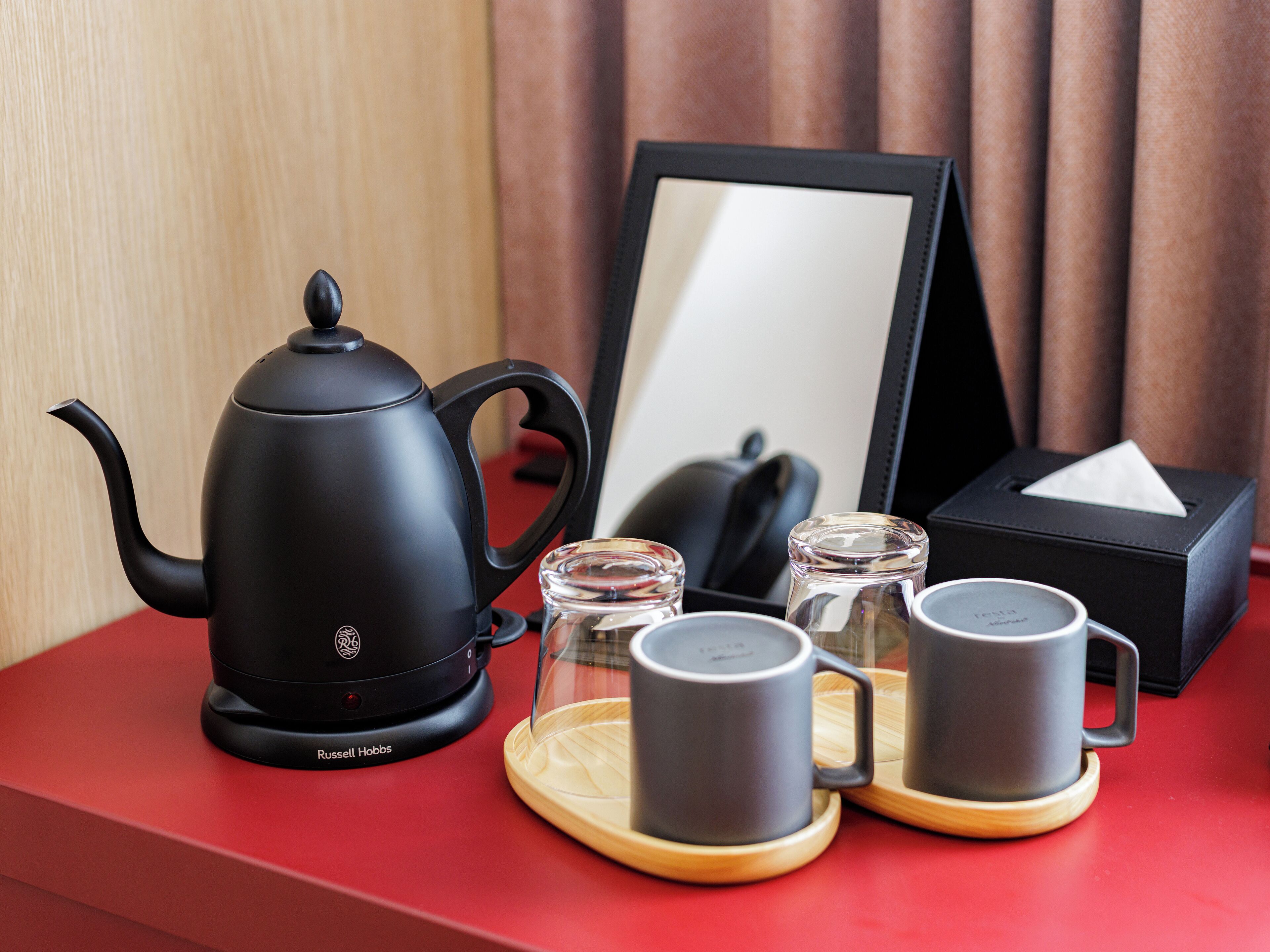 A black Russell Hobbs electric kettle, two clear glass mugs, and a black box with a tissue on a red table.