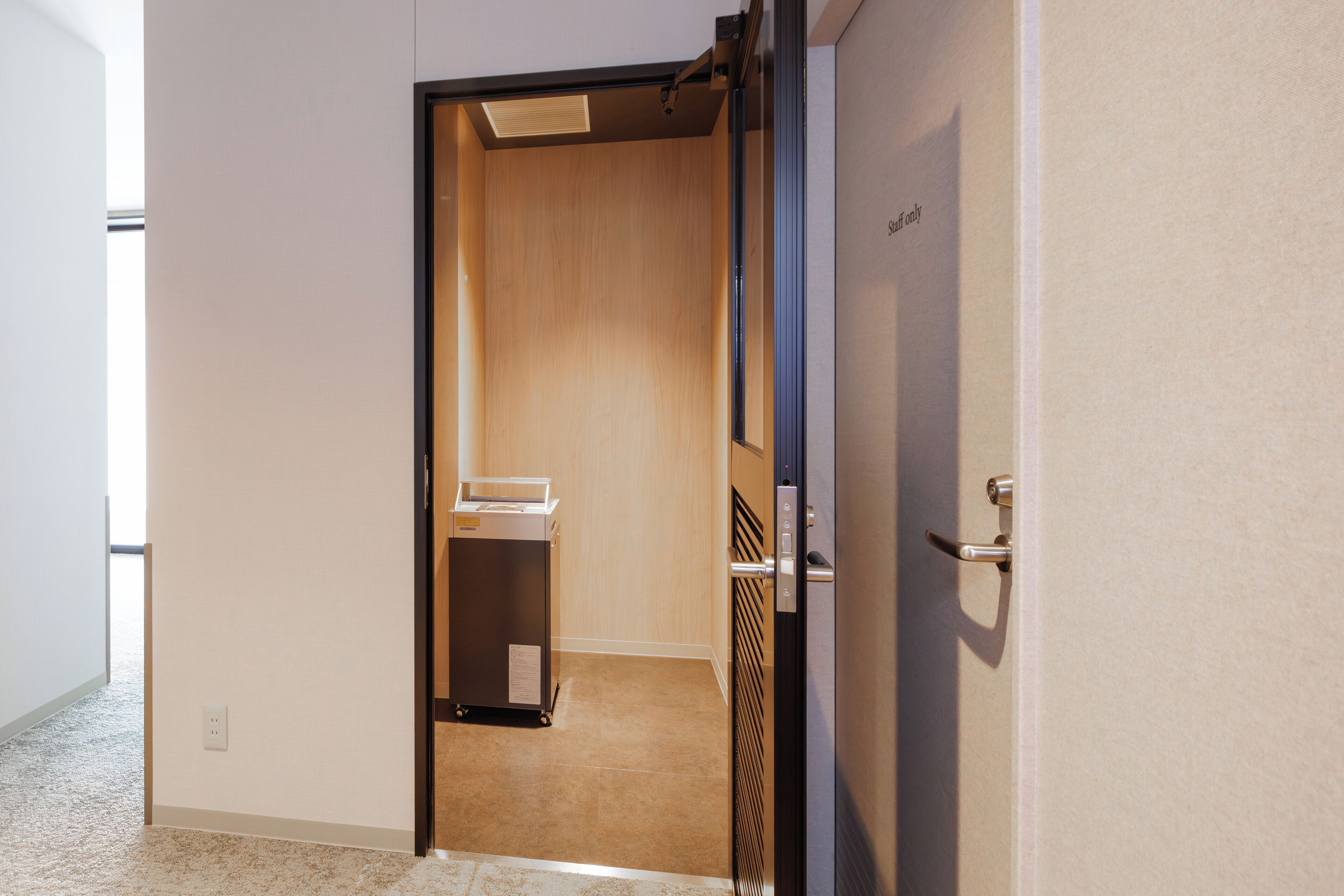 A hotel room with a wooden wardrobe, a small table, and a trash bin.
