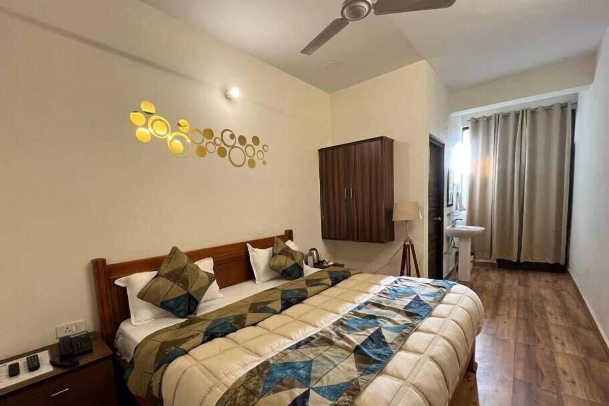 Una habitación de hotel con una cama, un ventilador de techo, una obra de arte en la pared, un armario de madera, una mesita de noche con una lámpara y un baño con un lavabo y cortinas.