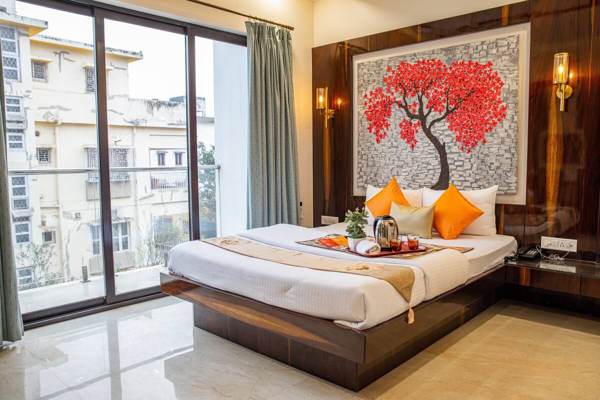 A hotel room with a large bed, a view of buildings through the window, and a wall-mounted painting of a tree.