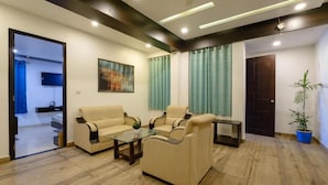 A living room with two beige sofas, a glass coffee table, and a small side table. There is a painting on the wall, a potted plant, and a door leading to another room. - Satkar Retreat (Udaipur)