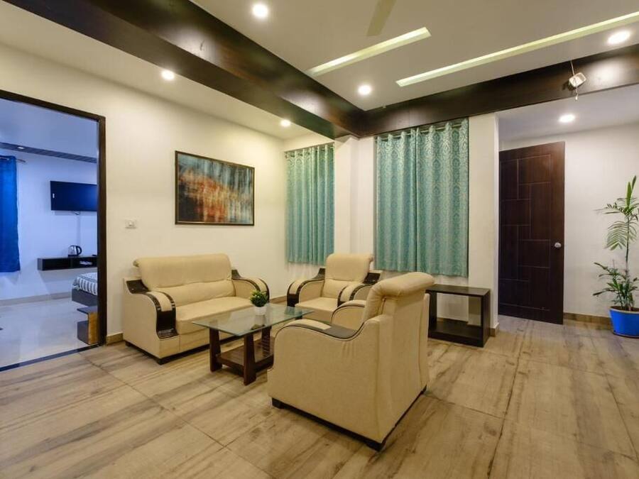 Una sala de estar con dos sofás color beige, una mesa de centro de vidrio y una mesita auxiliar. Hay un cuadro en la pared, una planta en maceta y una puerta que conduce a otra habitación.