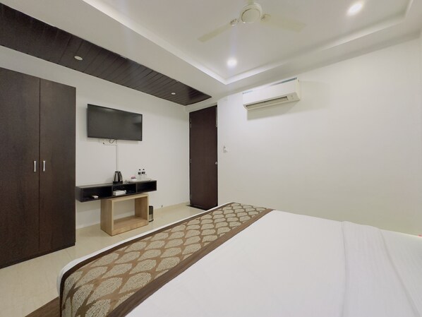 A hotel room with a bed, a TV mounted on the wall, a wardrobe, a bedside table with a kettle, and an air conditioning unit. - Satkar Retreat (Udaipur)