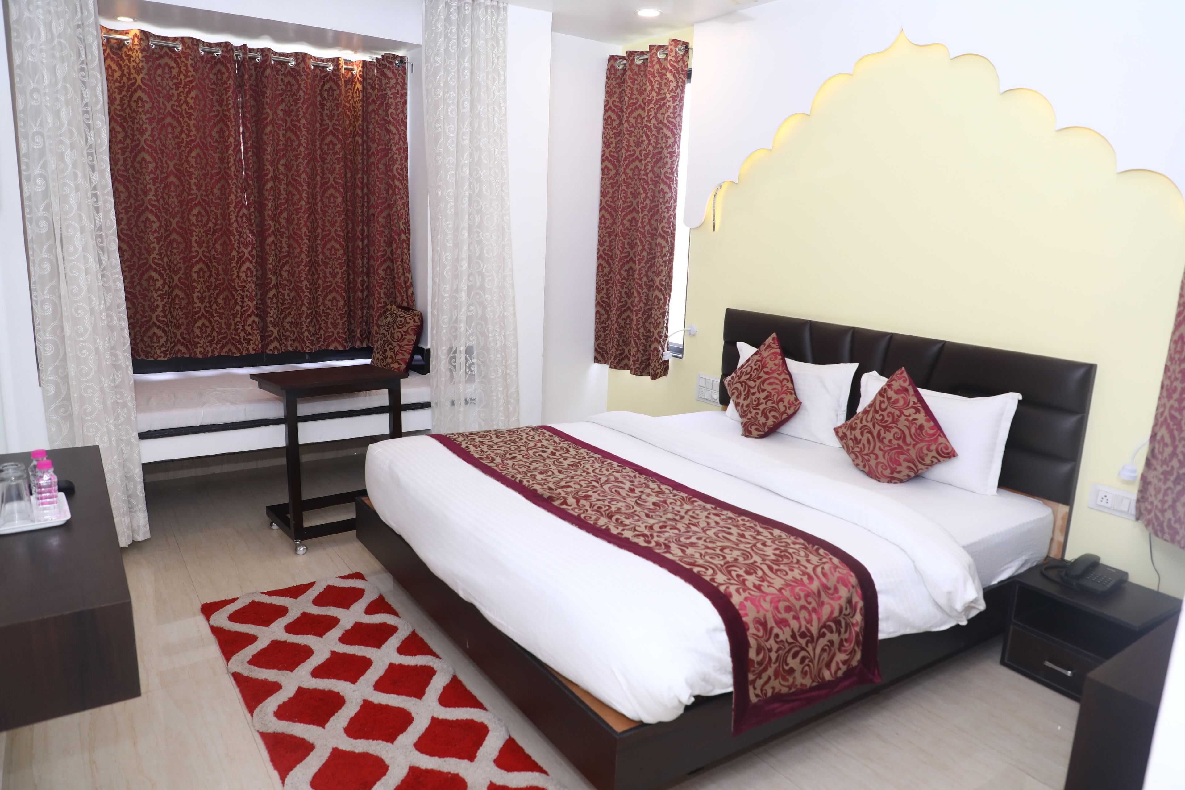 Une chambre d’hôtel avec un grand lit, une table de chevet, une commode, un tapis à motifs rouges et blancs et des rideaux à motifs.