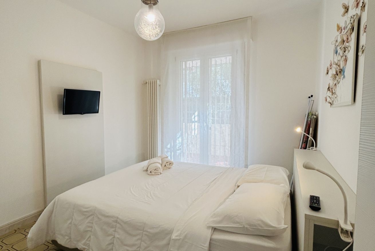 Une chambre à coucher comprenant un lit, un téléviseur fixé au mur, un radiateur, une fenêtre avec des rideaux légers et un luminaire fixé au mur.