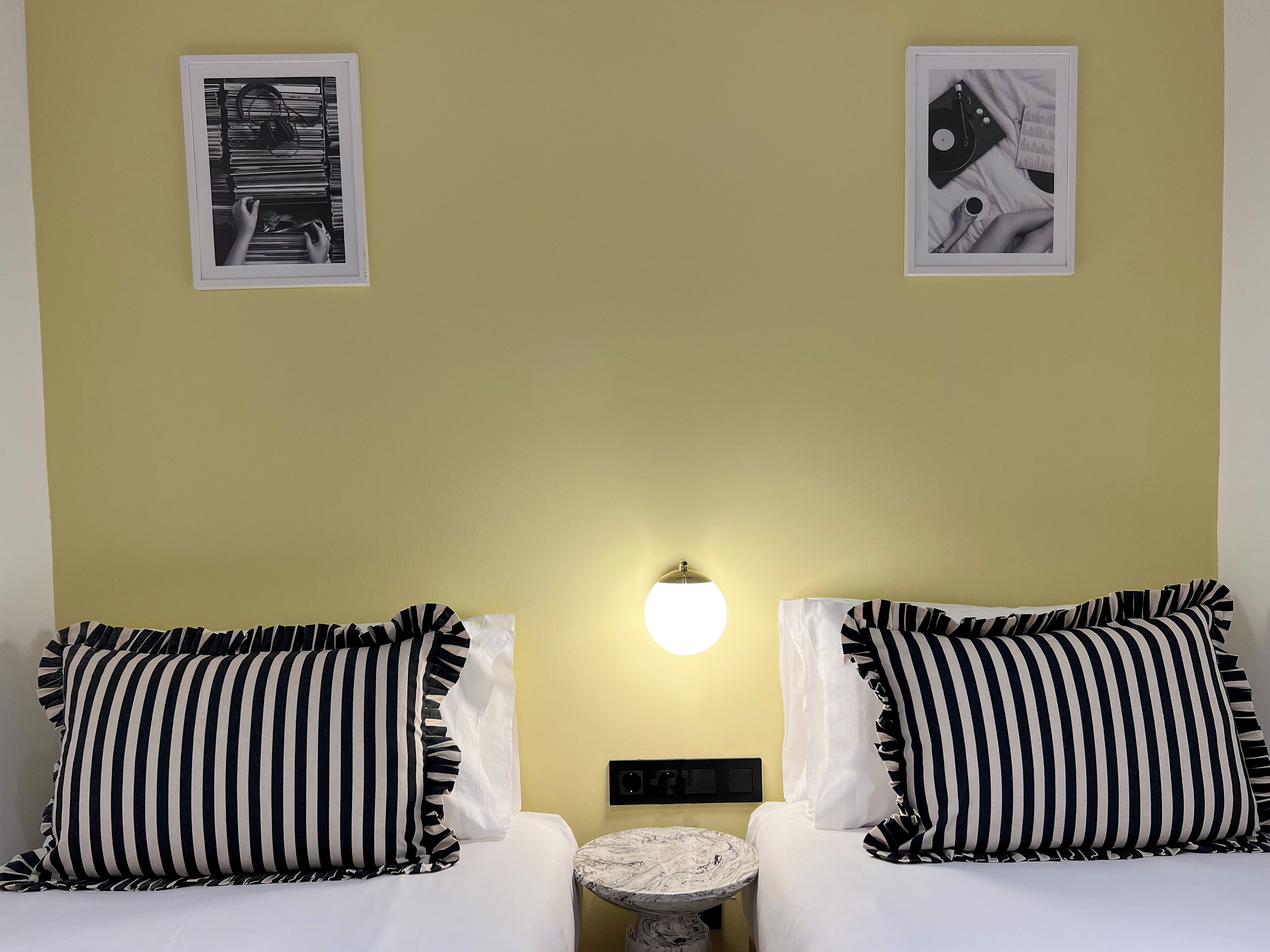 A room with two beds, each with black and white striped pillows, a small round table, and a wall-mounted light.
