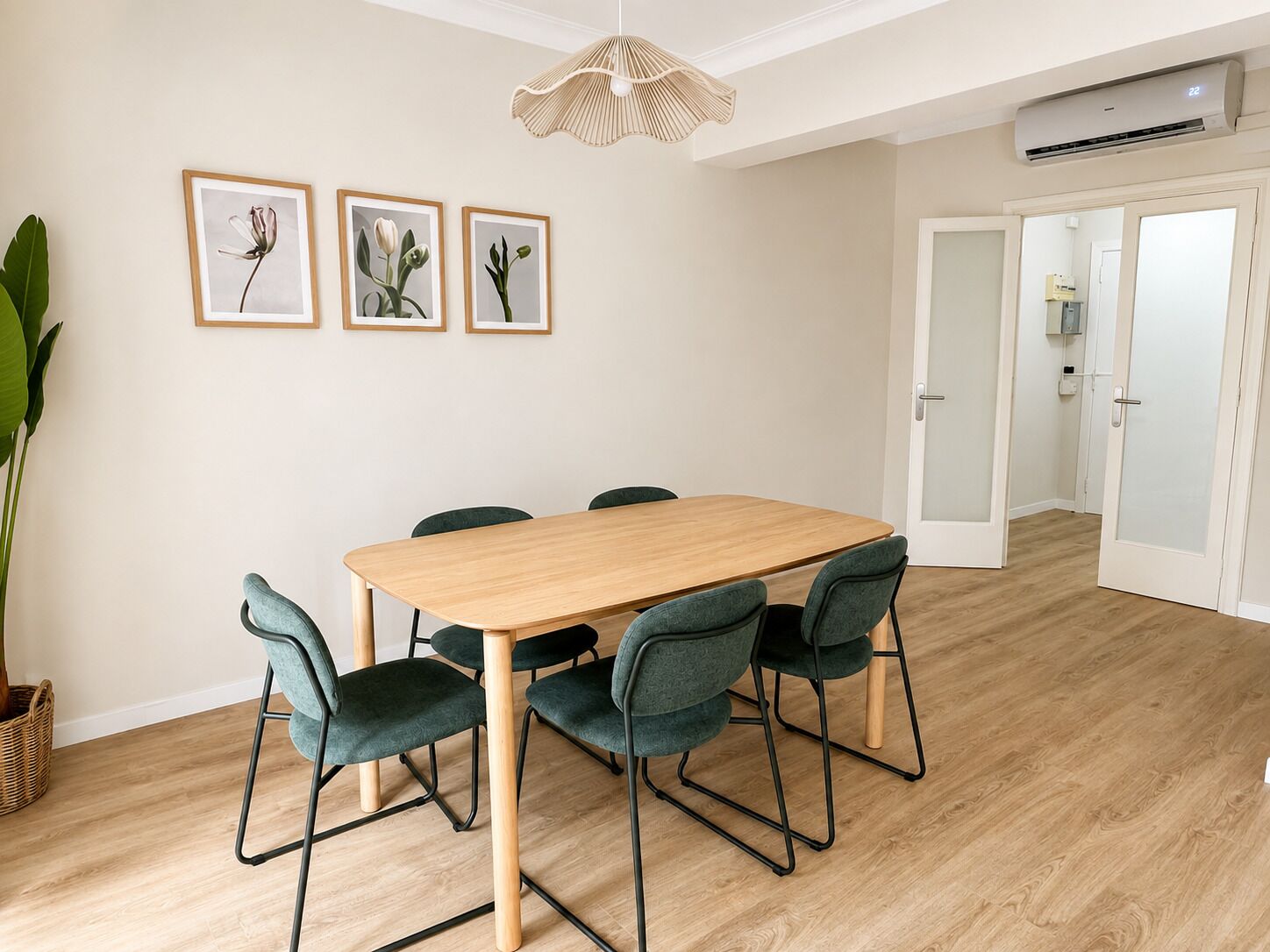 A dining area with a wooden table and green chairs, framed botanical prints on the wall, and a wicker pendant light.