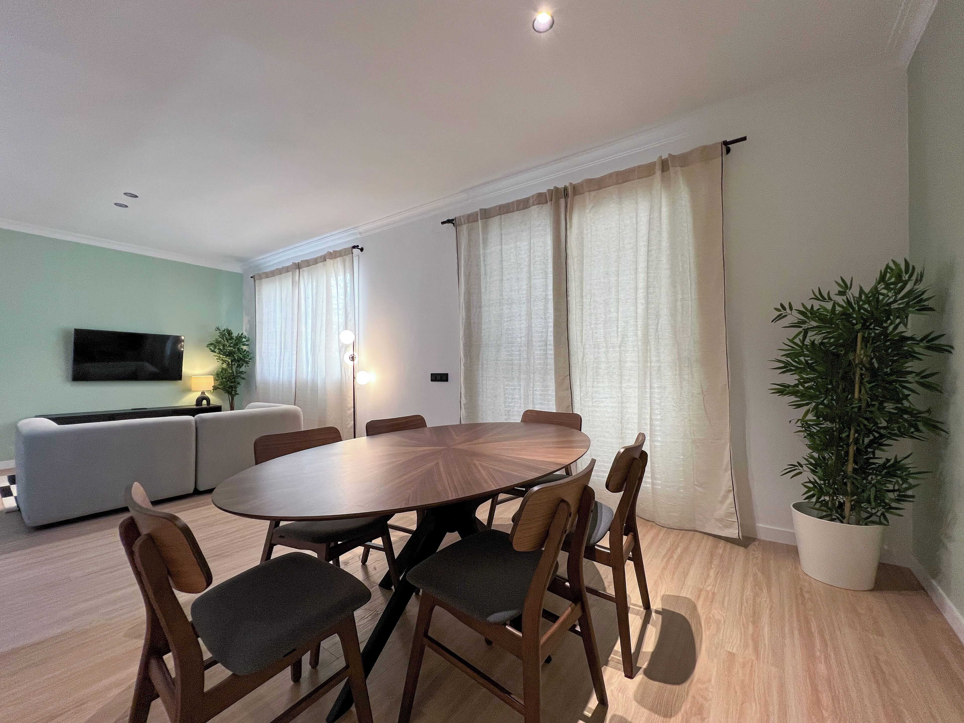 A modern living room with a round wooden dining table surrounded by chairs, a grey sofa, a flat-screen TV, and a potted plant.