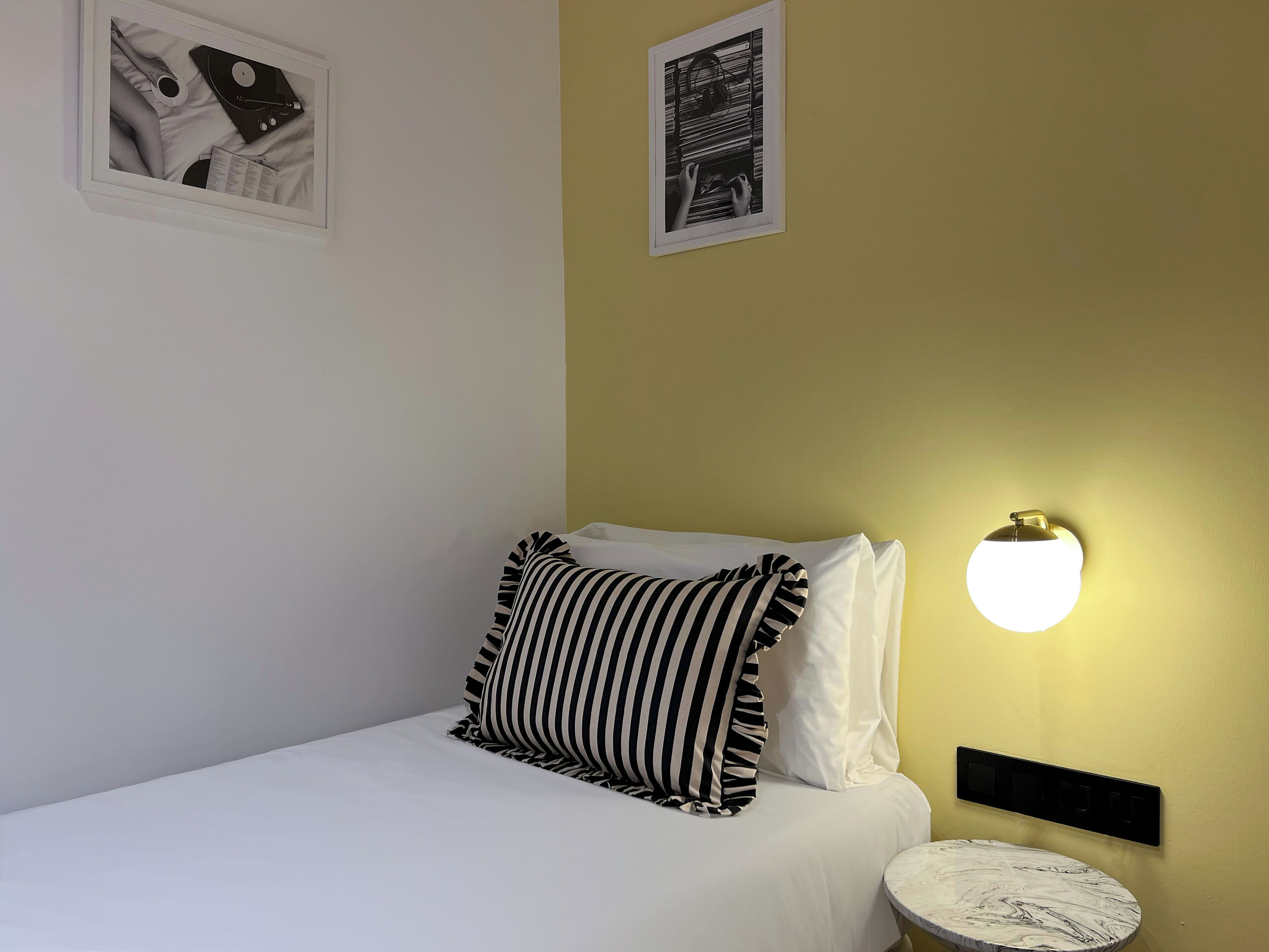 A neatly made bed with white bedding and striped pillows, a small round table with a marble top, and a wall-mounted light fixture.