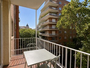 Een balkon met een tafel en stoelen, met uitzicht op een meerhoog gebouw met balkons en een boom.