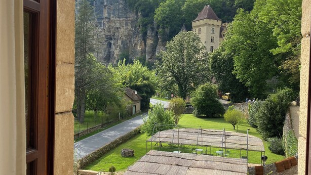 A view from a window overlooking a courtyard with a thatched-roof structure, a stone building, and a road.