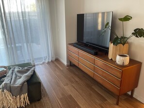 Een moderne woonkamer met een houten tv-meubel, een flatscreen televisie, een groene bank en een groot raam met dunne gordijnen.