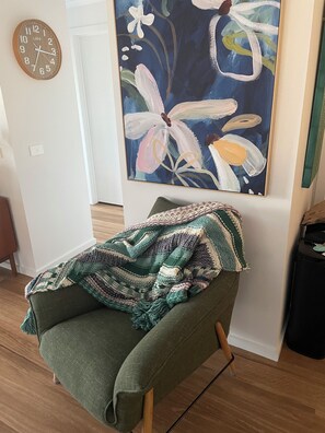 Een kamer met een groene fauteuil, een wandklok en een groot bloemen schilderij.