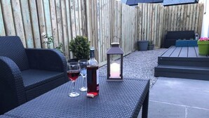 Een terras met een rieten tafel, twee stoelen, een lantaarn en een kleine potplant.