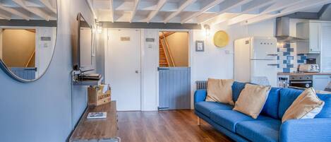 A compact living space with a blue sofa, wooden flooring, a kitchen area with a refrigerator, and a staircase leading to another level.