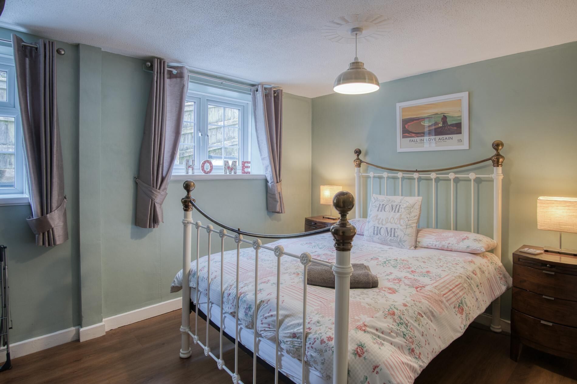 Une chambre à coucher avec un lit en métal blanc, une literie à motifs floraux, une table de chevet avec une lampe et une fenêtre avec des rideaux.