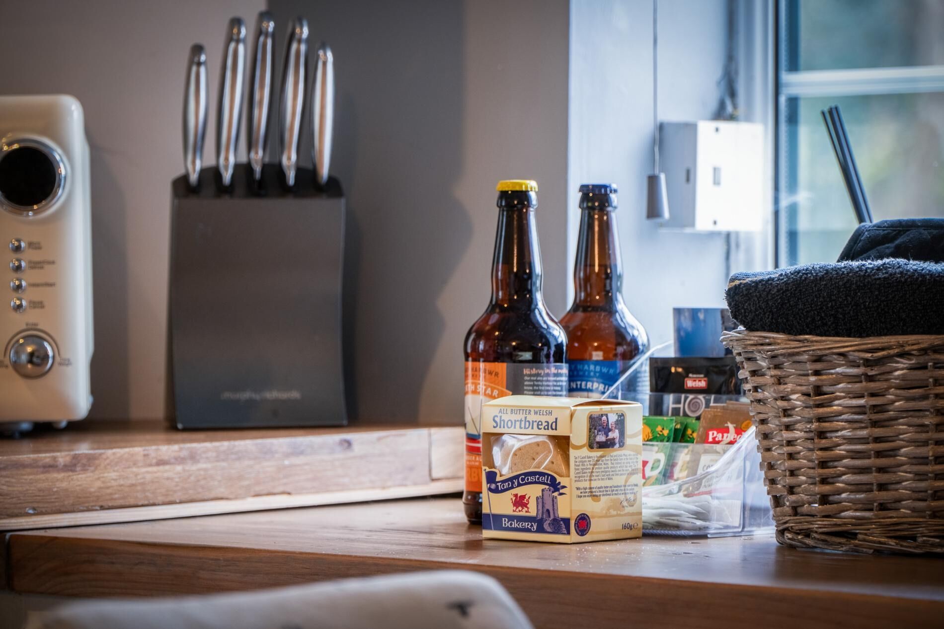 A kitchen counter with a bread box, two bottles of beer, and a wicker basket.
